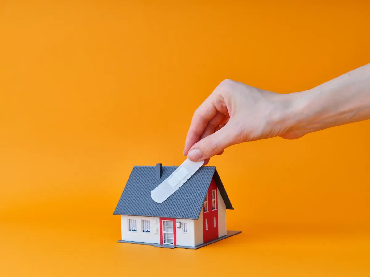Une main pose un pansement sur le toit d'une maquette de maison